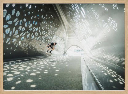 A skateboarder is captured mid-air performing a trick inside a futuristic, metallic tunnel with patterned cutouts that cast dappled light and shadows across the scene, all enclosed within a silver frame. Artwork