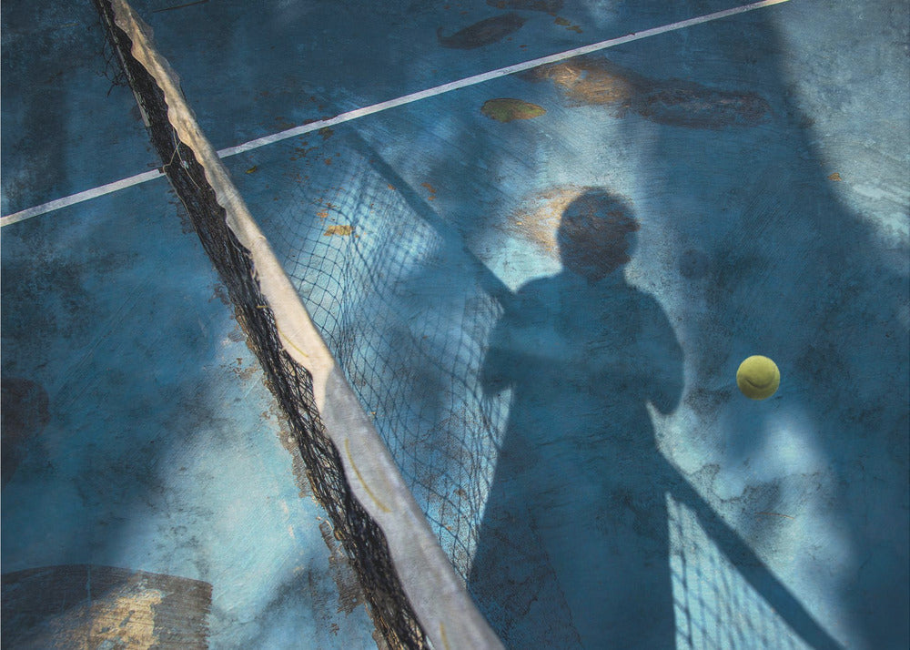 An artistic, high-angle shot of a weathered blue tennis court. The shadow of a tennis player holding a racket is cast across the court, with a yellow tennis ball suspended in mid-air nearby. A tattered tennis net runs diagonally across the frame, all enclosed within a silver border. Wall Art