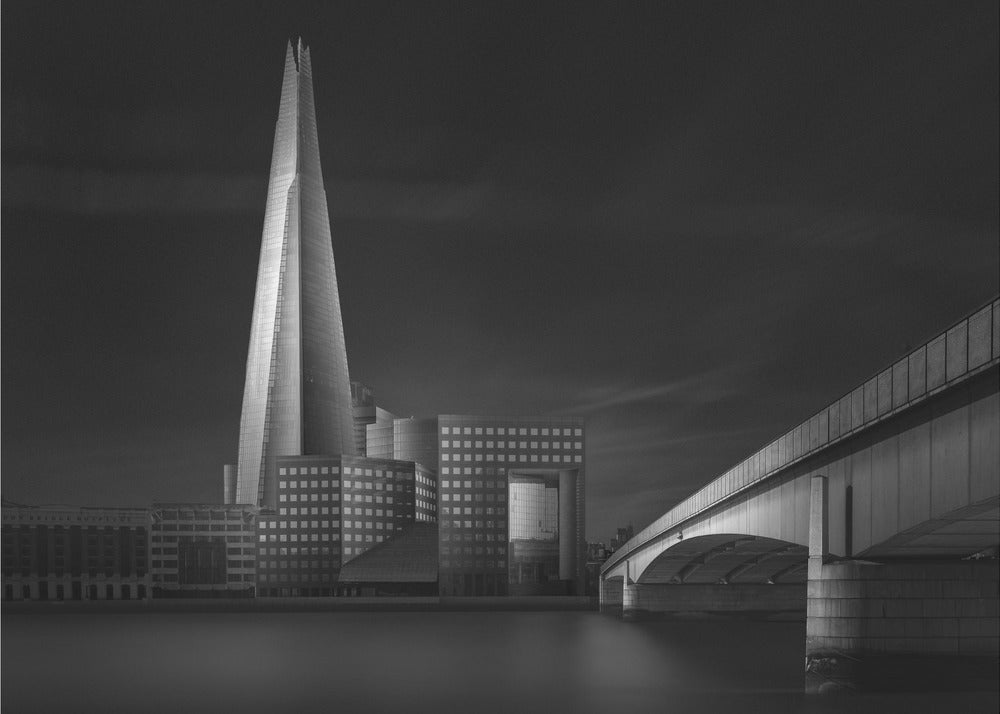 A dramatic black and white photograph of the London skyline featuring The Shard skyscraper and London Bridge. The image is shot at dusk or night, with The Shard brightly lit against a dark, moody sky. The River Thames in the foreground is calm and reflective, captured with a long exposure. Artwork