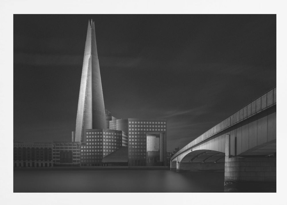 A dramatic black and white photograph of the London skyline featuring The Shard skyscraper and London Bridge. The image is shot at dusk or night, with The Shard brightly lit against a dark, moody sky. The River Thames in the foreground is calm and reflective, captured with a long exposure. Artwork
