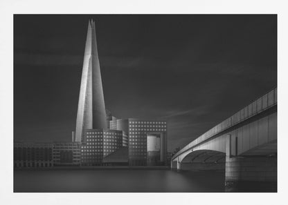 A dramatic black and white photograph of the London skyline featuring The Shard skyscraper and London Bridge. The image is shot at dusk or night, with The Shard brightly lit against a dark, moody sky. The River Thames in the foreground is calm and reflective, captured with a long exposure. Artwork