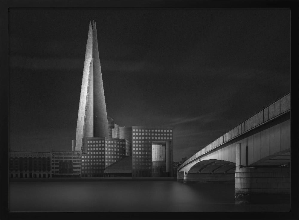 A dramatic black and white photograph of the London skyline featuring The Shard skyscraper and London Bridge. The image is shot at dusk or night, with The Shard brightly lit against a dark, moody sky. The River Thames in the foreground is calm and reflective, captured with a long exposure. Artwork
