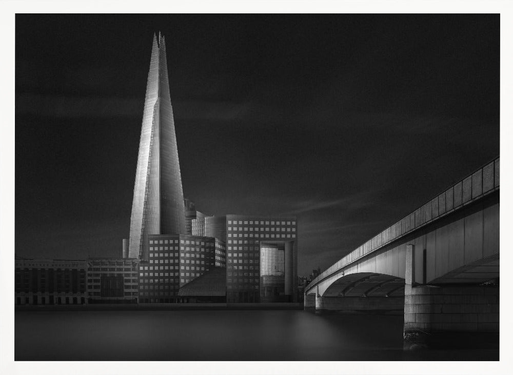 A dramatic black and white photograph of the London skyline featuring The Shard skyscraper and London Bridge. The image is shot at dusk or night, with The Shard brightly lit against a dark, moody sky. The River Thames in the foreground is calm and reflective, captured with a long exposure. Artwork