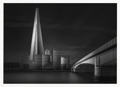 A dramatic black and white photograph of the London skyline featuring The Shard skyscraper and London Bridge. The image is shot at dusk or night, with The Shard brightly lit against a dark, moody sky. The River Thames in the foreground is calm and reflective, captured with a long exposure. Artwork
