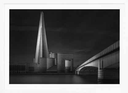 A dramatic black and white photograph of the London skyline featuring The Shard skyscraper and London Bridge. The image is shot at dusk or night, with The Shard brightly lit against a dark, moody sky. The River Thames in the foreground is calm and reflective, captured with a long exposure. Artwork