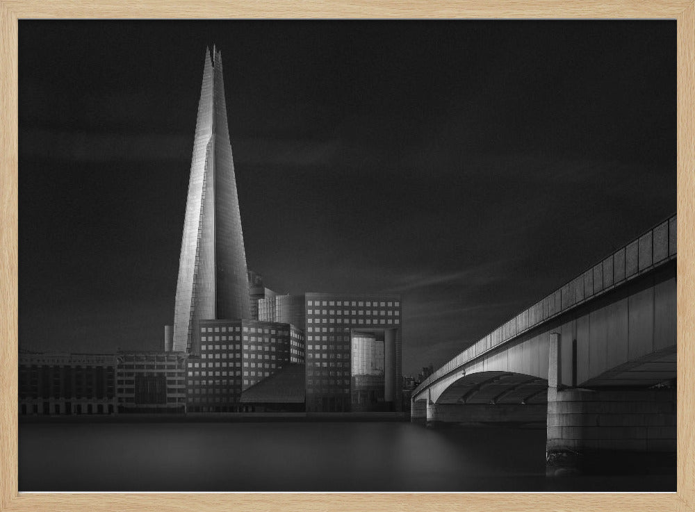 A dramatic black and white photograph of the London skyline featuring The Shard skyscraper and London Bridge. The image is shot at dusk or night, with The Shard brightly lit against a dark, moody sky. The River Thames in the foreground is calm and reflective, captured with a long exposure. Artwork