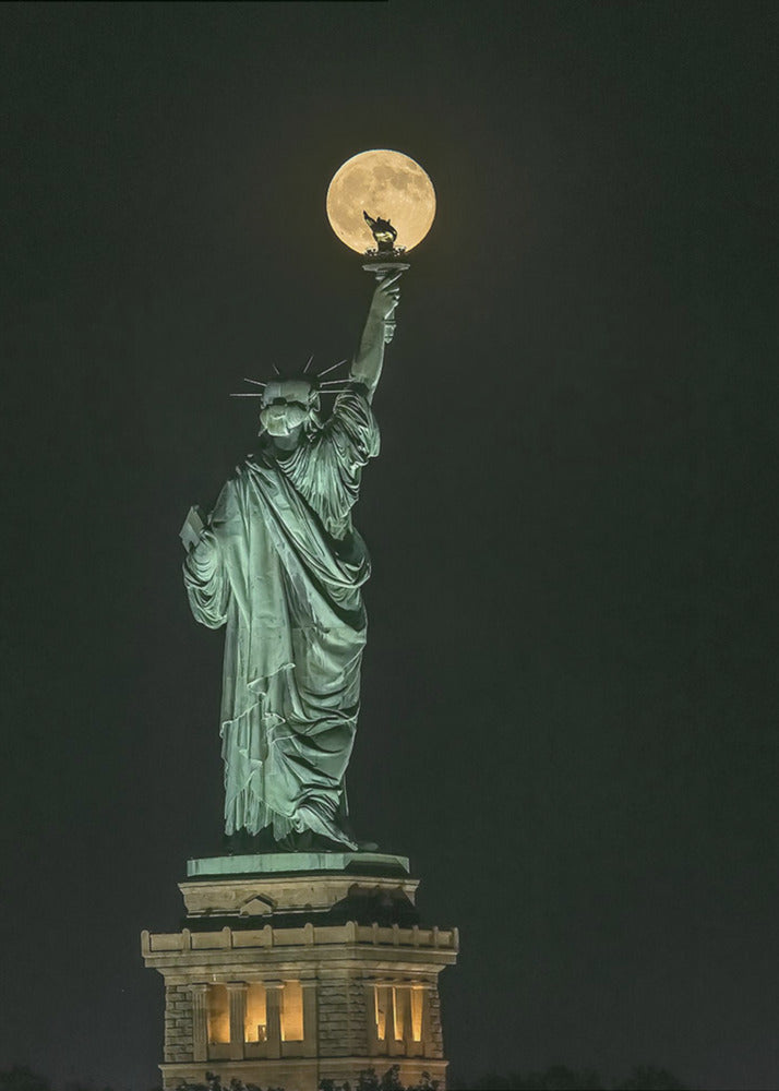 A striking night photograph of the Statue of Liberty, where a large, full yellow moon is perfectly aligned behind the torch, creating the illusion that the statue is holding up the moon. Artwork