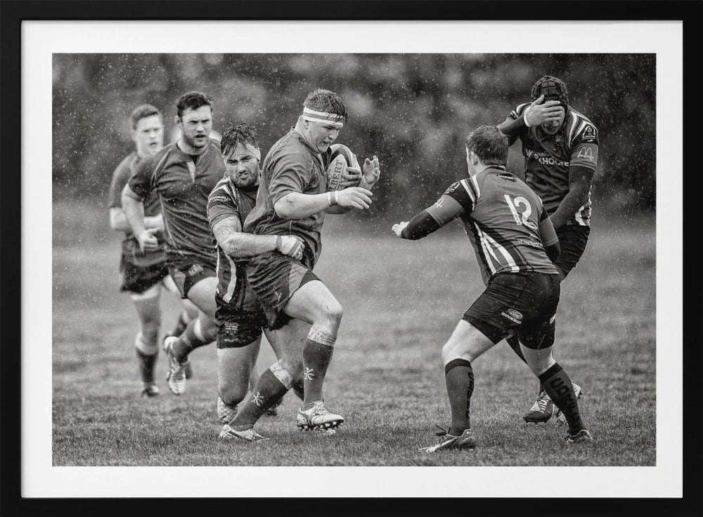 A dynamic black and white photograph of a rugby match being played in the rain. One player charges forward with the ball while an opponent wraps his arms around him in a tackle. Other players are in motion around them, capturing the intensity and physicality of the sport. Decor