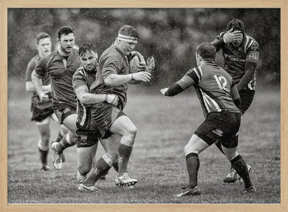 A dynamic black and white photograph of a rugby match being played in the rain. One player charges forward with the ball while an opponent wraps his arms around him in a tackle. Other players are in motion around them, capturing the intensity and physicality of the sport. Decor