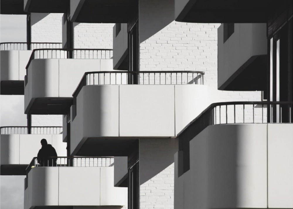 A black and white architectural photograph of a modern building with repeating geometric balconies. Strong sunlight creates deep shadows, emphasizing the lines and curves of the structure. A lone silhouetted figure stands on one of the lower balconies. Decor
