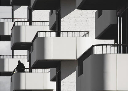 A black and white architectural photograph of a modern building with repeating geometric balconies. Strong sunlight creates deep shadows, emphasizing the lines and curves of the structure. A lone silhouetted figure stands on one of the lower balconies. Decor