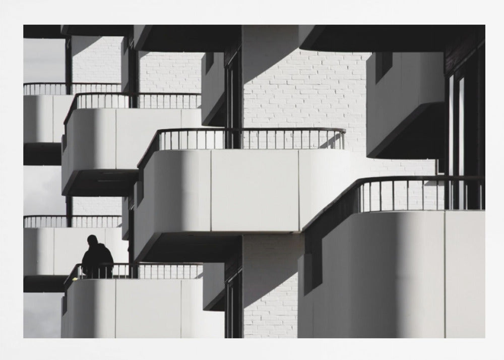 A black and white architectural photograph of a modern building with repeating geometric balconies. Strong sunlight creates deep shadows, emphasizing the lines and curves of the structure. A lone silhouetted figure stands on one of the lower balconies. Decor
