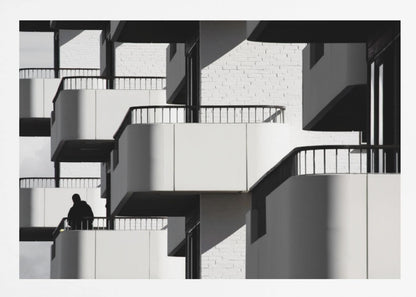 A black and white architectural photograph of a modern building with repeating geometric balconies. Strong sunlight creates deep shadows, emphasizing the lines and curves of the structure. A lone silhouetted figure stands on one of the lower balconies. Decor
