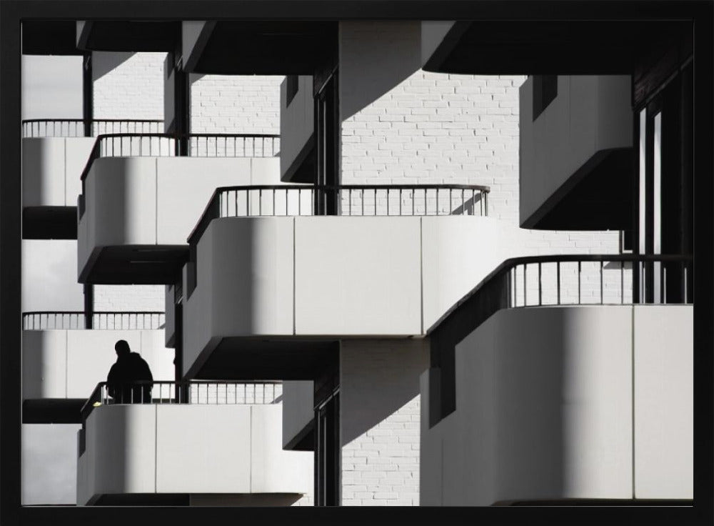 A black and white architectural photograph of a modern building with repeating geometric balconies. Strong sunlight creates deep shadows, emphasizing the lines and curves of the structure. A lone silhouetted figure stands on one of the lower balconies. Decor