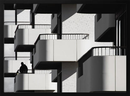 A black and white architectural photograph of a modern building with repeating geometric balconies. Strong sunlight creates deep shadows, emphasizing the lines and curves of the structure. A lone silhouetted figure stands on one of the lower balconies. Decor