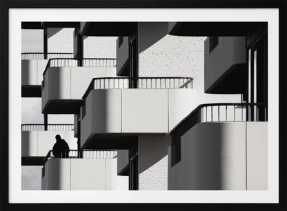 A black and white architectural photograph of a modern building with repeating geometric balconies. Strong sunlight creates deep shadows, emphasizing the lines and curves of the structure. A lone silhouetted figure stands on one of the lower balconies. Decor