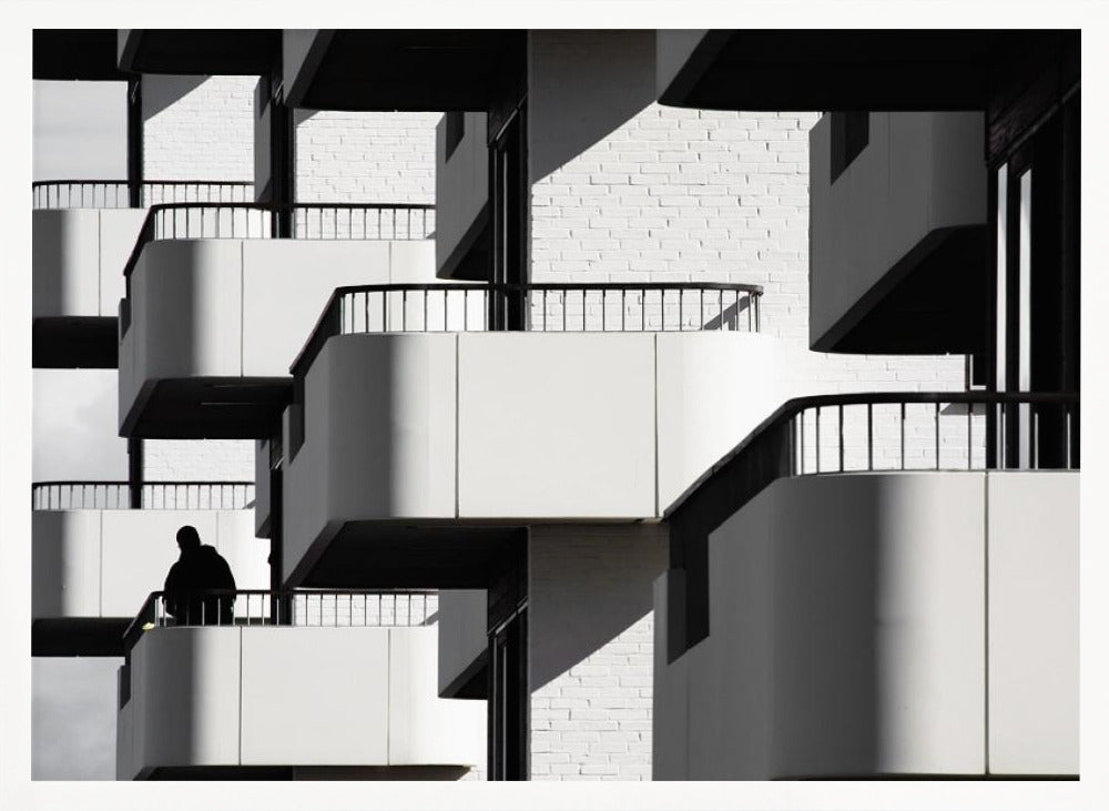 A black and white architectural photograph of a modern building with repeating geometric balconies. Strong sunlight creates deep shadows, emphasizing the lines and curves of the structure. A lone silhouetted figure stands on one of the lower balconies. Decor