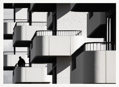 A black and white architectural photograph of a modern building with repeating geometric balconies. Strong sunlight creates deep shadows, emphasizing the lines and curves of the structure. A lone silhouetted figure stands on one of the lower balconies. Decor