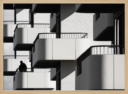 A black and white architectural photograph of a modern building with repeating geometric balconies. Strong sunlight creates deep shadows, emphasizing the lines and curves of the structure. A lone silhouetted figure stands on one of the lower balconies. Decor