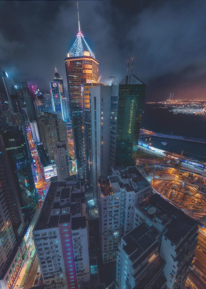 A dramatic high-angle nighttime photograph of a sprawling modern cityscape, with towering skyscrapers illuminated in vibrant blue, orange, and purple neon lights under a dark, cloudy sky. Below, streets are aglow with traffic, and in the distance, a brightly lit harbor meets the dark water. Decor
