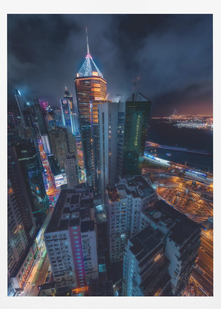 A dramatic high-angle nighttime photograph of a sprawling modern cityscape, with towering skyscrapers illuminated in vibrant blue, orange, and purple neon lights under a dark, cloudy sky. Below, streets are aglow with traffic, and in the distance, a brightly lit harbor meets the dark water. Decor