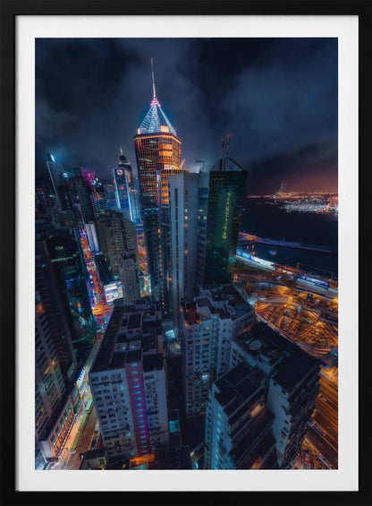 A dramatic high-angle nighttime photograph of a sprawling modern cityscape, with towering skyscrapers illuminated in vibrant blue, orange, and purple neon lights under a dark, cloudy sky. Below, streets are aglow with traffic, and in the distance, a brightly lit harbor meets the dark water. Decor