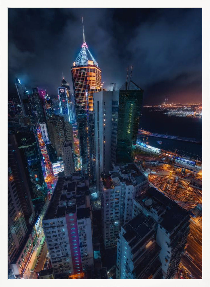 A dramatic high-angle nighttime photograph of a sprawling modern cityscape, with towering skyscrapers illuminated in vibrant blue, orange, and purple neon lights under a dark, cloudy sky. Below, streets are aglow with traffic, and in the distance, a brightly lit harbor meets the dark water. Decor