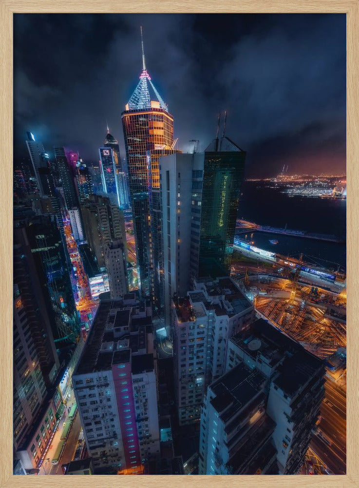 A dramatic high-angle nighttime photograph of a sprawling modern cityscape, with towering skyscrapers illuminated in vibrant blue, orange, and purple neon lights under a dark, cloudy sky. Below, streets are aglow with traffic, and in the distance, a brightly lit harbor meets the dark water. Decor