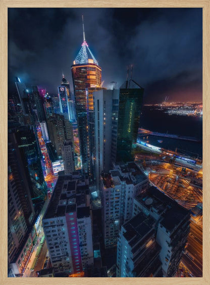 A dramatic high-angle nighttime photograph of a sprawling modern cityscape, with towering skyscrapers illuminated in vibrant blue, orange, and purple neon lights under a dark, cloudy sky. Below, streets are aglow with traffic, and in the distance, a brightly lit harbor meets the dark water. Decor