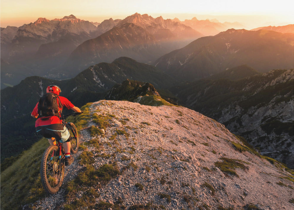 A mountain biker wearing a red shirt and helmet rides along a narrow, rocky mountain ridge at sunset. The background features a vast landscape of layered mountain ranges glowing in the warm, golden light of the setting sun. Poster