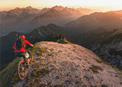 A mountain biker wearing a red shirt and helmet rides along a narrow, rocky mountain ridge at sunset. The background features a vast landscape of layered mountain ranges glowing in the warm, golden light of the setting sun. Poster