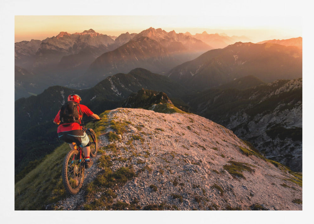 A mountain biker wearing a red shirt and helmet rides along a narrow, rocky mountain ridge at sunset. The background features a vast landscape of layered mountain ranges glowing in the warm, golden light of the setting sun. Poster