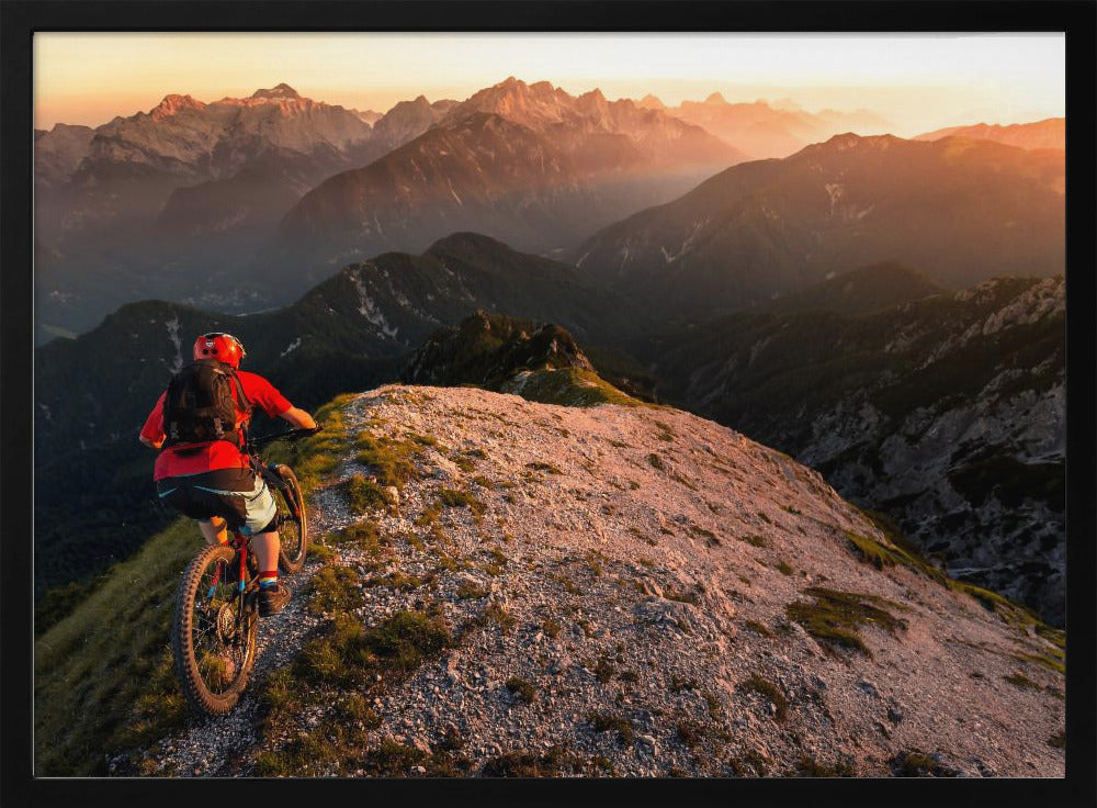 A mountain biker wearing a red shirt and helmet rides along a narrow, rocky mountain ridge at sunset. The background features a vast landscape of layered mountain ranges glowing in the warm, golden light of the setting sun. Poster