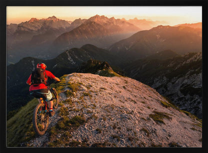 A mountain biker wearing a red shirt and helmet rides along a narrow, rocky mountain ridge at sunset. The background features a vast landscape of layered mountain ranges glowing in the warm, golden light of the setting sun. Poster