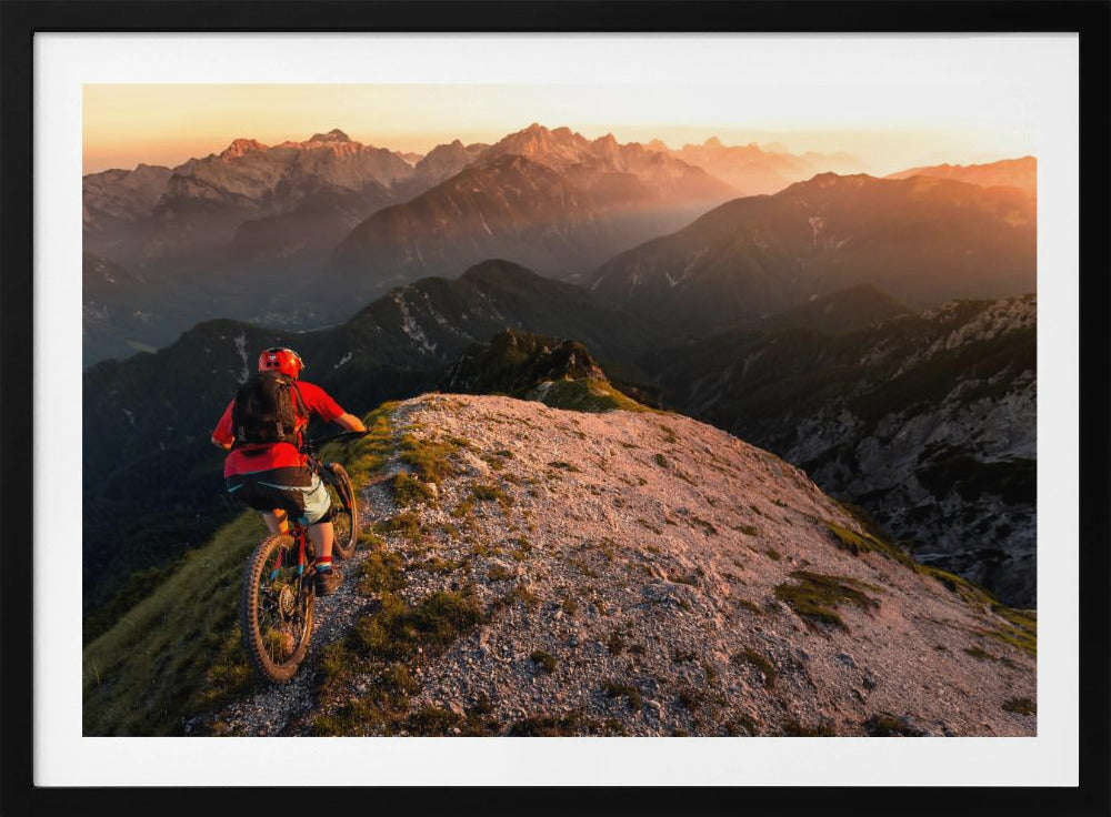 A mountain biker wearing a red shirt and helmet rides along a narrow, rocky mountain ridge at sunset. The background features a vast landscape of layered mountain ranges glowing in the warm, golden light of the setting sun. Poster