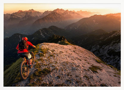 A mountain biker wearing a red shirt and helmet rides along a narrow, rocky mountain ridge at sunset. The background features a vast landscape of layered mountain ranges glowing in the warm, golden light of the setting sun. Poster
