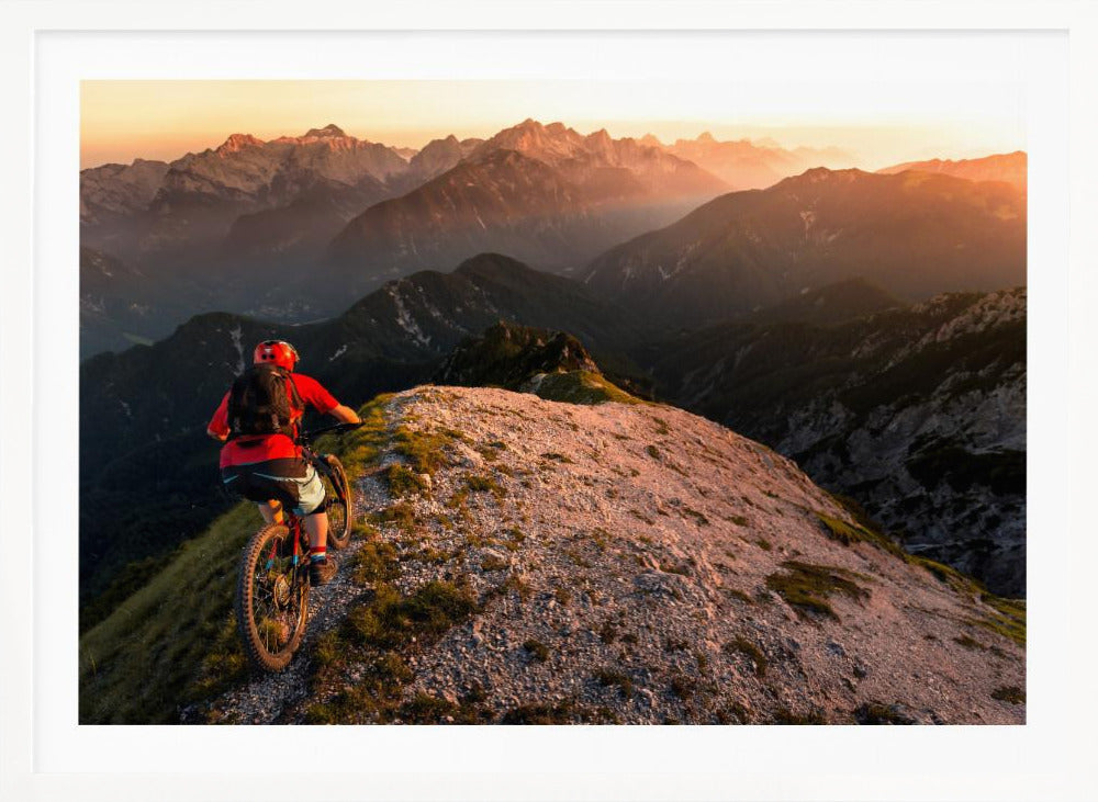A mountain biker wearing a red shirt and helmet rides along a narrow, rocky mountain ridge at sunset. The background features a vast landscape of layered mountain ranges glowing in the warm, golden light of the setting sun. Poster