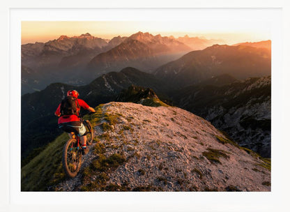 A mountain biker wearing a red shirt and helmet rides along a narrow, rocky mountain ridge at sunset. The background features a vast landscape of layered mountain ranges glowing in the warm, golden light of the setting sun. Poster