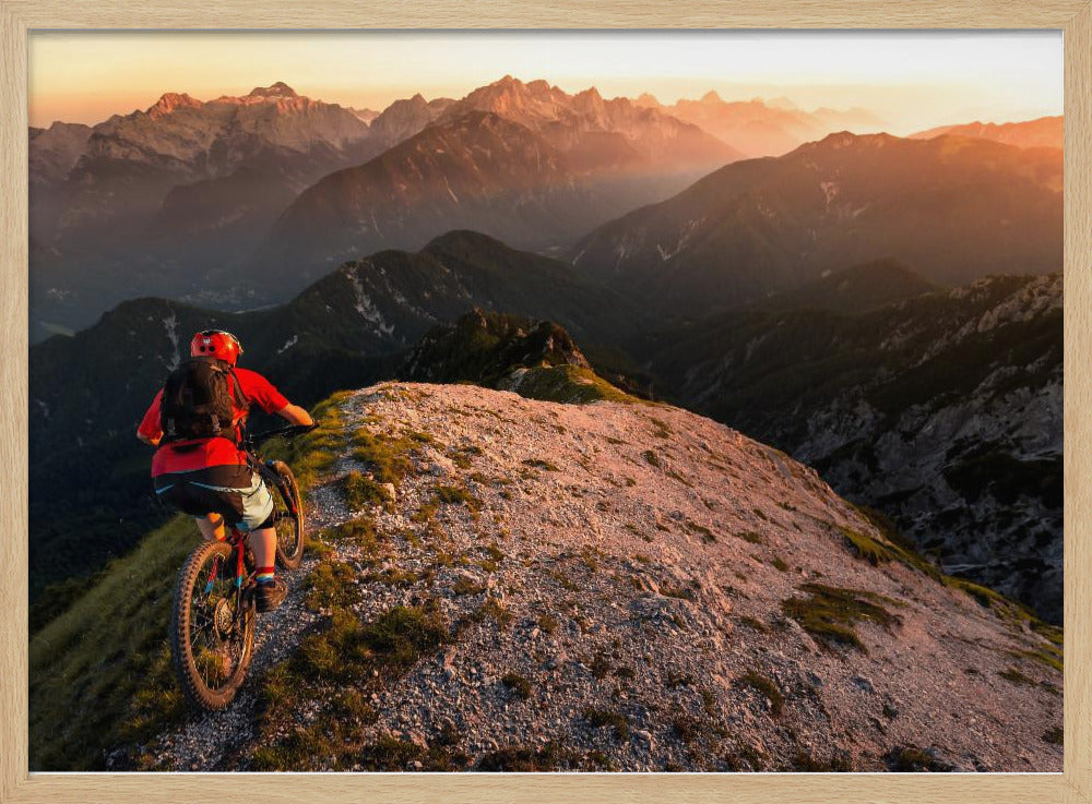 A mountain biker wearing a red shirt and helmet rides along a narrow, rocky mountain ridge at sunset. The background features a vast landscape of layered mountain ranges glowing in the warm, golden light of the setting sun. Poster