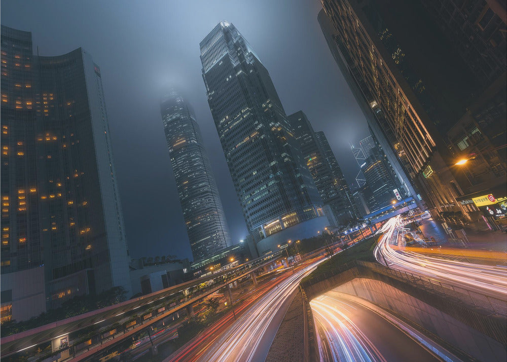 A framed, low-angle photograph capturing a bustling city at night, with towering skyscrapers shrouded in fog. Long-exposure creates vibrant streaks of light from traffic on a multi-level highway, illuminating the dark, modern urban landscape. Wall Art