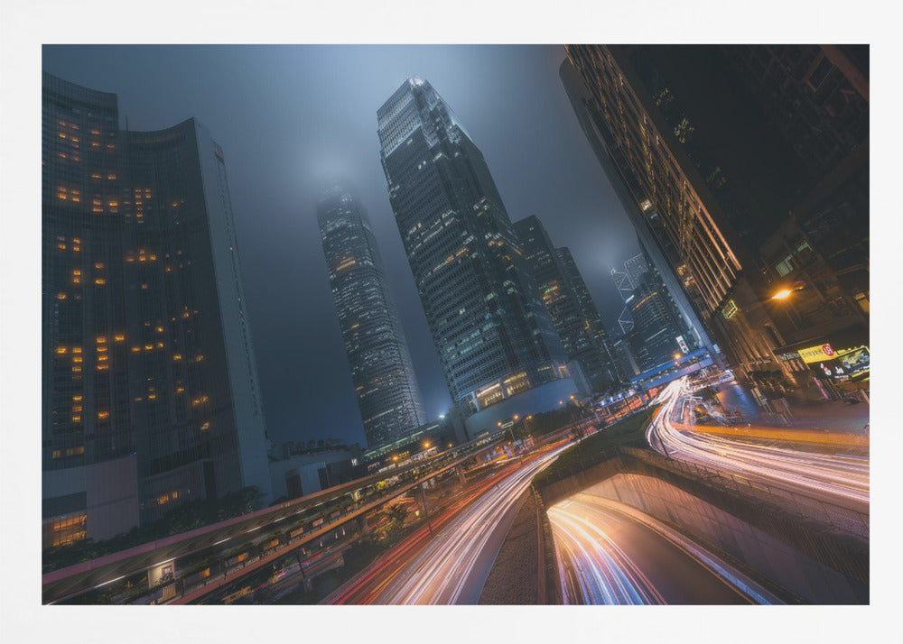 A framed, low-angle photograph capturing a bustling city at night, with towering skyscrapers shrouded in fog. Long-exposure creates vibrant streaks of light from traffic on a multi-level highway, illuminating the dark, modern urban landscape. Wall Art