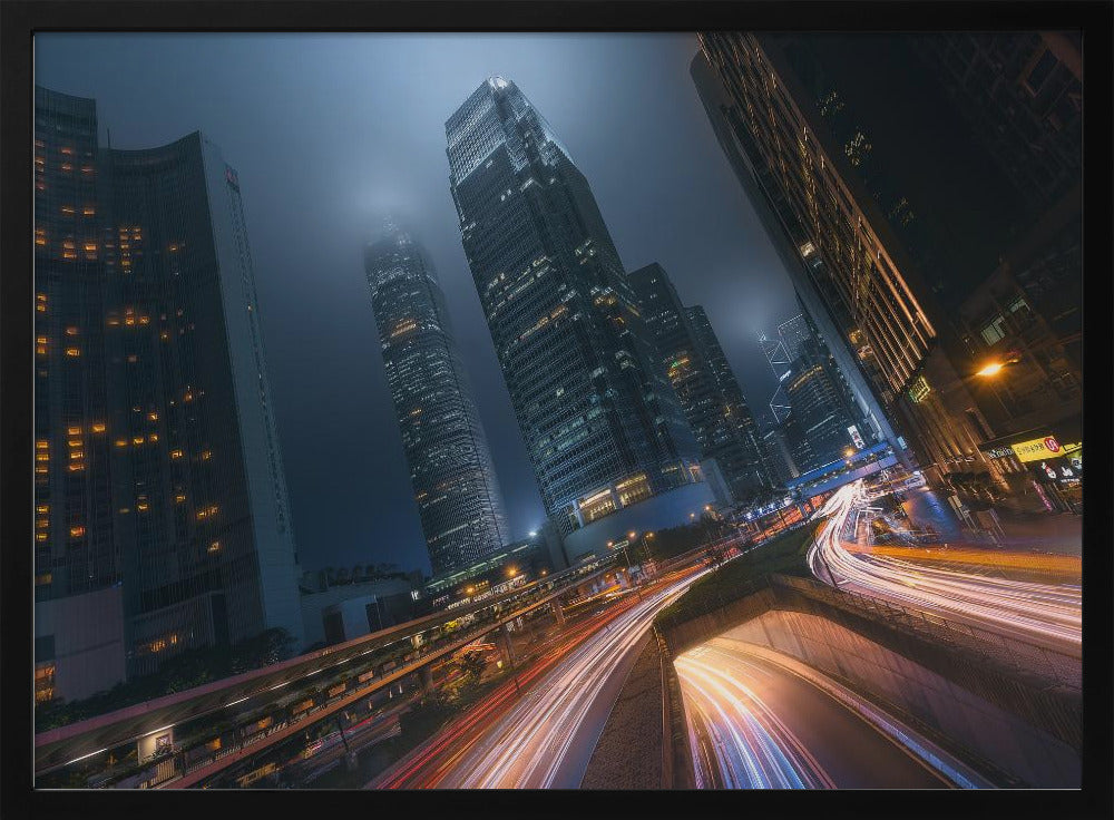 A framed, low-angle photograph capturing a bustling city at night, with towering skyscrapers shrouded in fog. Long-exposure creates vibrant streaks of light from traffic on a multi-level highway, illuminating the dark, modern urban landscape. Wall Art