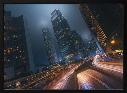 A framed, low-angle photograph capturing a bustling city at night, with towering skyscrapers shrouded in fog. Long-exposure creates vibrant streaks of light from traffic on a multi-level highway, illuminating the dark, modern urban landscape. Wall Art