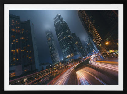 A framed, low-angle photograph capturing a bustling city at night, with towering skyscrapers shrouded in fog. Long-exposure creates vibrant streaks of light from traffic on a multi-level highway, illuminating the dark, modern urban landscape. Wall Art