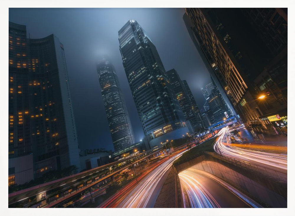 A framed, low-angle photograph capturing a bustling city at night, with towering skyscrapers shrouded in fog. Long-exposure creates vibrant streaks of light from traffic on a multi-level highway, illuminating the dark, modern urban landscape. Wall Art