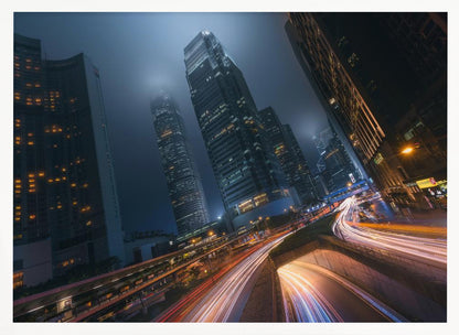 A framed, low-angle photograph capturing a bustling city at night, with towering skyscrapers shrouded in fog. Long-exposure creates vibrant streaks of light from traffic on a multi-level highway, illuminating the dark, modern urban landscape. Wall Art
