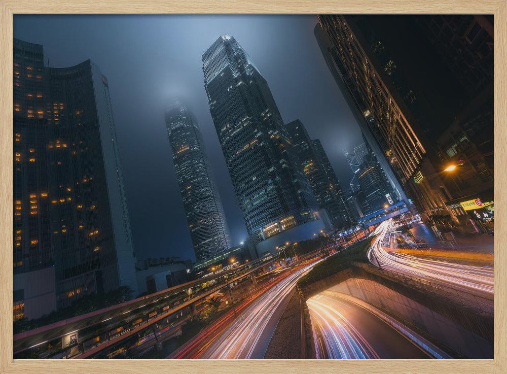 A framed, low-angle photograph capturing a bustling city at night, with towering skyscrapers shrouded in fog. Long-exposure creates vibrant streaks of light from traffic on a multi-level highway, illuminating the dark, modern urban landscape. Wall Art