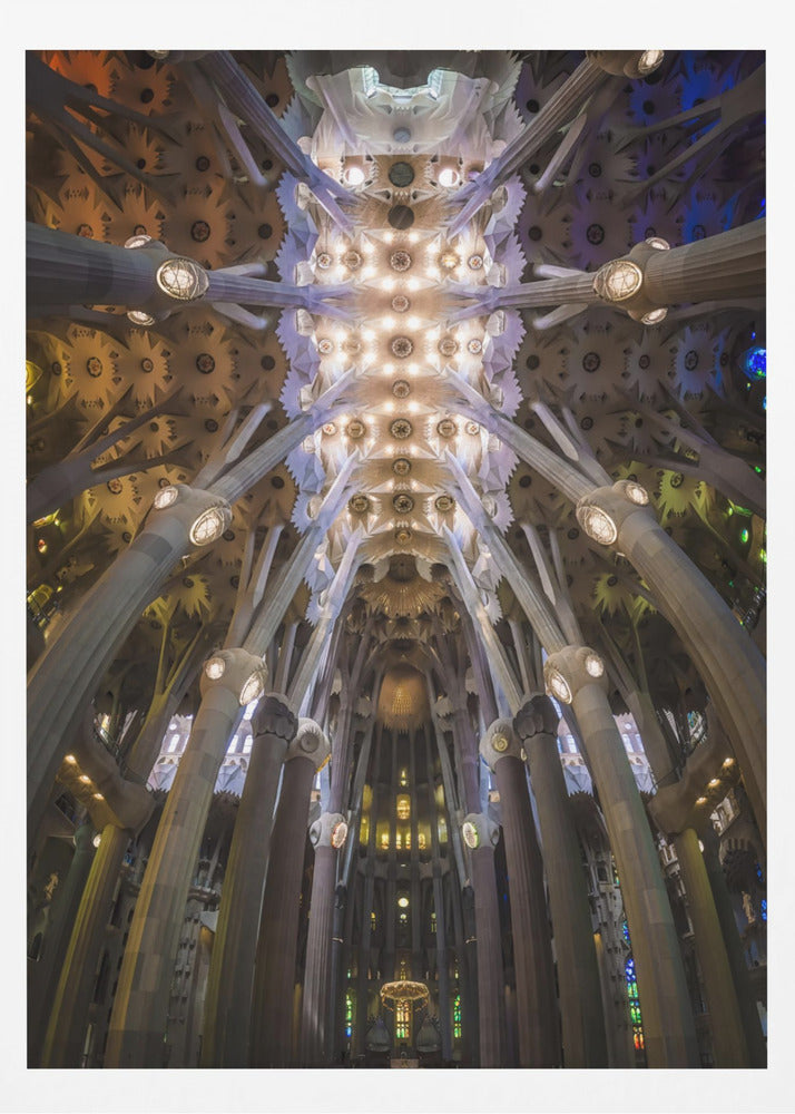 A low-angle, symmetrical photograph looking up at the incredibly ornate and complex vaulted ceiling of the Sagrada Familia Basilica. The massive stone columns branch out like trees, supporting the intricately detailed ceiling illuminated by warm, glowing lights and splashes of color from unseen stained glass windows. Poster