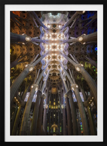 A low-angle, symmetrical photograph looking up at the incredibly ornate and complex vaulted ceiling of the Sagrada Familia Basilica. The massive stone columns branch out like trees, supporting the intricately detailed ceiling illuminated by warm, glowing lights and splashes of color from unseen stained glass windows. Poster