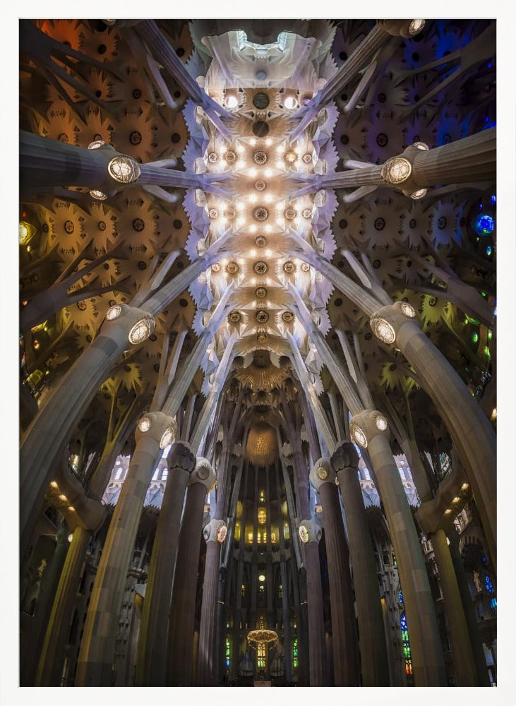 A low-angle, symmetrical photograph looking up at the incredibly ornate and complex vaulted ceiling of the Sagrada Familia Basilica. The massive stone columns branch out like trees, supporting the intricately detailed ceiling illuminated by warm, glowing lights and splashes of color from unseen stained glass windows. Poster